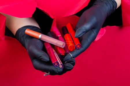 Photo for Gloved hands holding pile of various lip gloss sticks. Breaking through a red paper wall. - Royalty Free Image