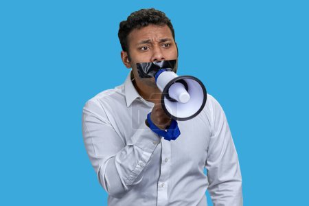Foto de Un joven indio con la boca pegada sostiene el megáfono. Aislado en azul. - Imagen libre de derechos