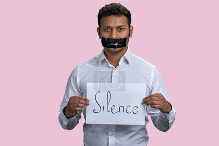 Foto de Hombre indio marrón sosteniendo hoja de papel con palabra de silencio. Aislado sobre fondo rosa. - Imagen libre de derechos