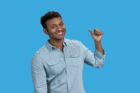 Photo for Young smiling south asian man is pointing back with thumb. Isolated on blue. - Royalty Free Image