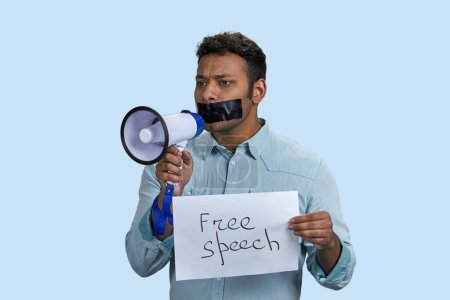 Photo for Young southasian man holding sheet of paper and megaphone. Indian guy with mouth taped. Isolated on blue background. - Royalty Free Image