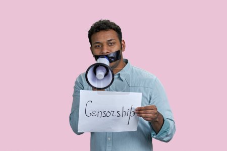 Foto de Joven hombre marrón con la boca pegada sosteniendo el megáfono y el papel con la palabra censura. Aislado sobre fondo rosa. - Imagen libre de derechos