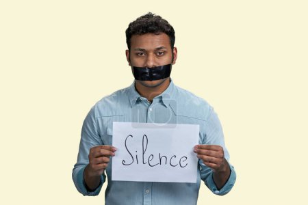 Photo for Indian man with mouth taped holding paper sheet with silence inscription. Isolated on beige background. - Royalty Free Image
