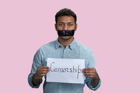 Téléchargez les photos : Un Indien réduit au silence avec une bouche collée tenant une feuille de papier avec un mot de censure. Isolé sur fond rose. - en image libre de droit