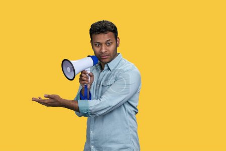 Téléchargez les photos : Jeune homme indien parlant en mégaphone et montrant quelque chose avec sa main. Isolé sur fond jaune vif. - en image libre de droit