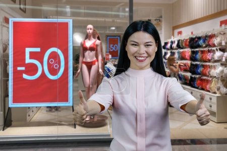 Photo for Happy young asian woman shows both thumb up. Lingerie boutique with half discount in the background. - Royalty Free Image