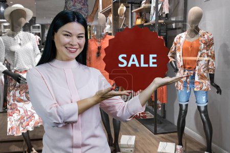 Photo for Smiling brunette asian woman advertising clothing shop discount. Boutique in the background. - Royalty Free Image