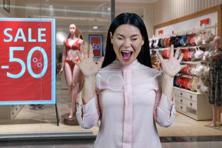 Photo for Young hysterical asian woman screaming out loud. Lingerie boutique with half discount in the background. - Royalty Free Image