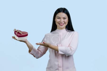 Téléchargez les photos : Portrait d'une femme asiatique souriante annonçant boîte-cadeau en forme de coeur. Isolé sur fond bleu. - en image libre de droit