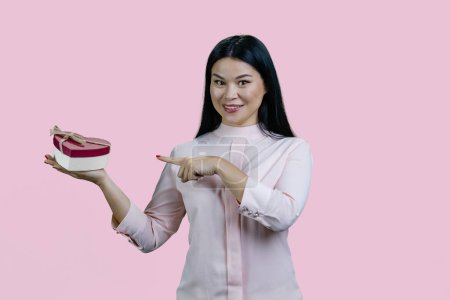 Téléchargez les photos : Portrait d'une jeune femme asiatique souriante pointant vers une boîte-cadeau en forme de cœur. Isolé sur fond rose. - en image libre de droit