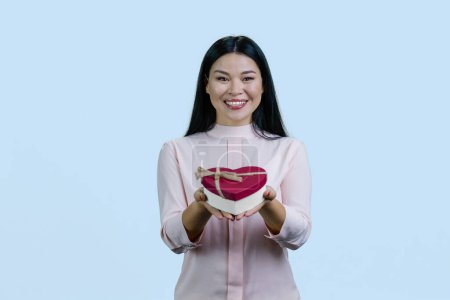 Téléchargez les photos : Portrait de heureuse une jeune femme asiatique gaie vous donnant une boîte cadeau en forme de coeur. Isolé sur fond bleu. - en image libre de droit