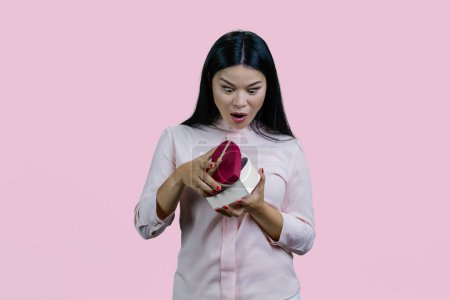 Téléchargez les photos : Heureuse jeune femme asiatique ouvre une boîte cadeau en forme de coeur. Étonnement et surprise avec les yeux grands ouverts. Isolé sur fond rose. - en image libre de droit