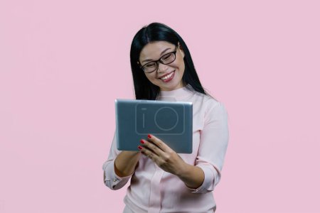 Téléchargez les photos : Gai sourire asiatique femme dans lunettes tenant comprimé pc. Isolé sur fond rose. - en image libre de droit