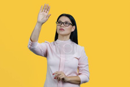 Foto de Joven mujer de negocios asiática en gafas es tocar pantalla invencible por su mano. Aislado sobre amarillo. - Imagen libre de derechos