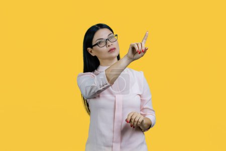 Photo for Young asian businesswoman is touching an invsible screen by her index finger. Pointing at emptiness for copy space. Isolated on yellow. - Royalty Free Image