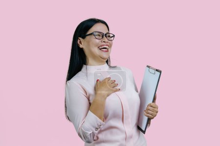 Foto de Joven mujer de negocios asiática en gafas sujetando portapapeles y riendo muy duro. Aislado sobre fondo rosa. - Imagen libre de derechos