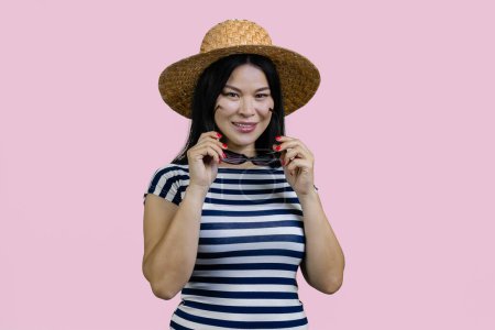 Foto de Retrato de una joven turista asiática en sombrero de paja se pone gafas de sol. Aislado sobre fondo rosa. - Imagen libre de derechos