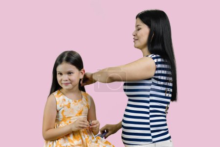 Téléchargez les photos : Jeune brune mère peigne les cheveux de sa petite fille. Isolé sur fond rose. - en image libre de droit