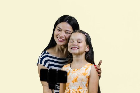 Téléchargez les photos : Mère et fille font un selfie sur smartphone à l'aide du bâton. Isolé sur jaune pastel. - en image libre de droit