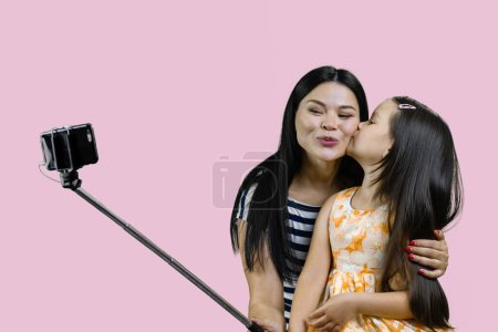 Téléchargez les photos : Mère et sa fille font un selfie sur smartphone à l'aide du bâton. Petite fille embrassant sa mère pour la photo. Isolé sur rose. - en image libre de droit