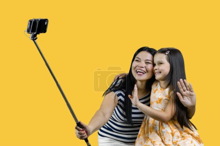 Téléchargez les photos : Maman et sa fille font une photo commune sur smartphone à l'aide d'un selfie stick. Isolé sur fond jaune. - en image libre de droit