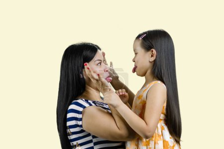 Téléchargez les photos : Vue de côté de la femme et la petite fille font des visages les uns aux autres. Isolé sur fond jaune pastel. - en image libre de droit