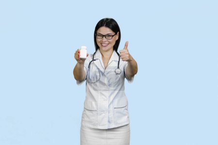 Foto de La doctora asiática con frasco de medicina blanca le está dando el pulgar hacia arriba. Aislado en azul pálido. - Imagen libre de derechos