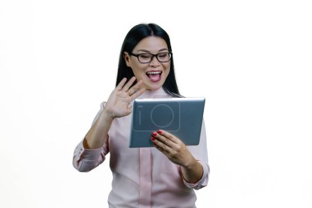 Téléchargez les photos : Jeune femme d'affaires asiatique tient une tablette et agite la main. Saluer quelqu'un en ligne via une conversation vidéo. Isolé sur fond blanc. - en image libre de droit