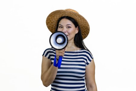 Téléchargez les photos : Jeune femme asiatique dans un chapeau de paille de bord annonce quelque chose en mégaphone. Isolé sur blanc. - en image libre de droit
