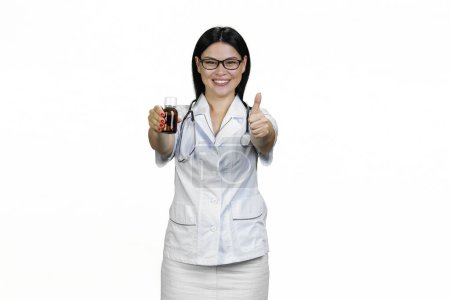 Foto de Retrato de mujer asiática sonriente médico con frasco de jarabe dando un pulgar hacia arriba. Aislado sobre blanco. - Imagen libre de derechos