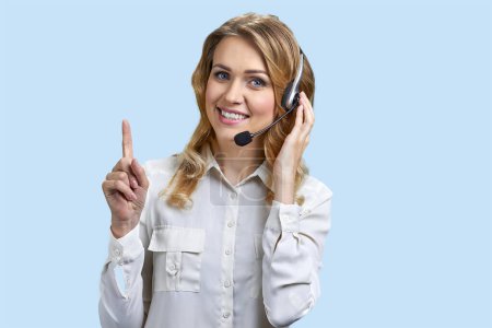 Photo for Smiling blond businesswoman with headset is pointing up. Pastel blue background. - Royalty Free Image