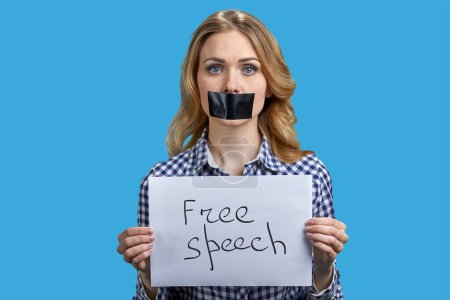 Foto de Joven mujer rubia manifestante con la boca pegada sosteniendo pancarta con la inscripción libertad de expresión. Mujer silenciada sobre fondo azul. - Imagen libre de derechos