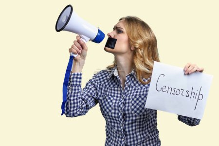 Foto de Mujer rubia joven con boca grabada sosteniendo megáfono y pancarta con inscripción de censura. Aislado sobre fondo amarillo. - Imagen libre de derechos