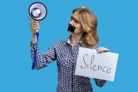 Foto de Mujer rubia con boca grabada sosteniendo megáfono y pancarta con inscripción de silencio. Aislado sobre fondo azul vivo. - Imagen libre de derechos