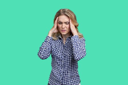 Photo for Portrait of young blond woman touching head because of headache. Stress and frustration. Isolated on green. - Royalty Free Image