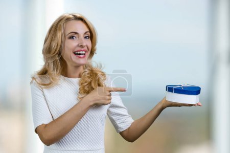 Foto de Mujer rubia de mediana edad positiva apuntando a la caja de regalo de forma de corazón azul en su mano. Fondo interior borroso abstracto. - Imagen libre de derechos