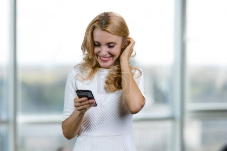 Téléchargez les photos : Heureuse femme d'affaires blonde souriante tenant son smartphone debout à l'intérieur. Fenêtre floue en arrière-plan. - en image libre de droit