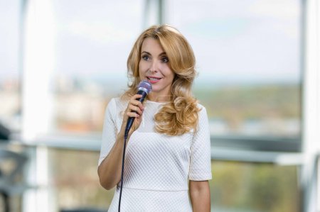 Photo for Portrait of blonde middle aged lady giving a speech in microphone. Blurred indoor window in the background. - Royalty Free Image