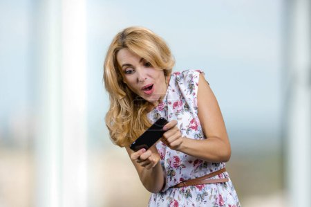 Photo for Excited blonde woman plays video game on her smartphone. Blurred window in the background. - Royalty Free Image