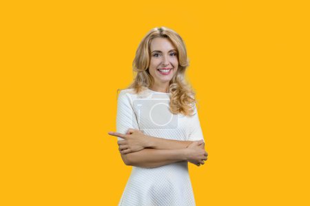 Téléchargez les photos : Portrait de heureuse femme blonde souriante avec les bras croisés pointant vers la droite. Isolé sur jaune. - en image libre de droit