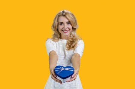 Photo for Happy caucasian blonde woman holding blue heart shape gift box. Isolated orange background. Giving a present to you. - Royalty Free Image