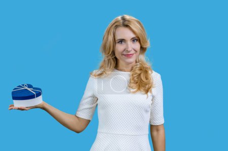 Photo for Happy mature blonde woman holding blue heart shape gift box on her right palm. Isolated blue background. Giving a present to you. - Royalty Free Image