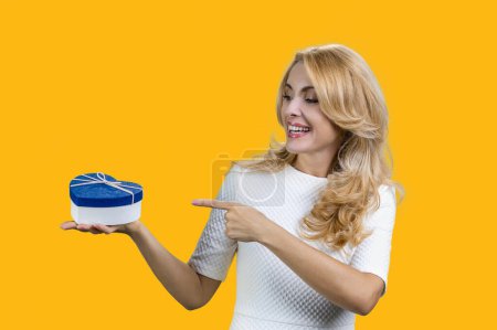 Photo for Happy blonde woman holding blue heart shape gift box and pointing with finger. Isolated on yellow. - Royalty Free Image