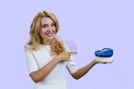 Photo for Happy blondie holding a blue heart shape gift box and pointing with index finger. Isolated on purple. - Royalty Free Image