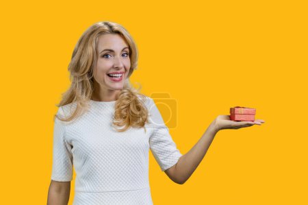 Photo for Happy mature blonde woman holding red gift box on her left palm. Isolated on yellow. - Royalty Free Image