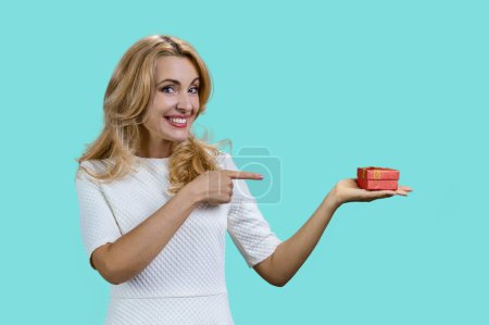 Photo for Happy mature blonde holding a red gift box and pointing with index finger. Isolated on turquoise. - Royalty Free Image