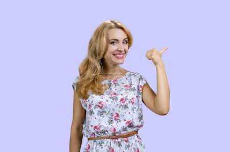 Photo for Portrait of attractive blonde mature woman pointing back with her thumb. Isolated on purple. - Royalty Free Image