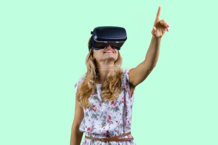 Photo for Happy surprised mature woman in virtual reality headset pointing up. Isolated on turquoise. - Royalty Free Image