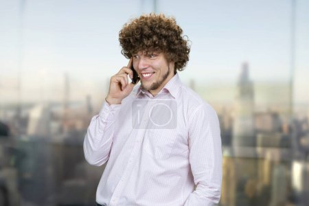 Téléchargez les photos : Portrait d'un jeune caucasien en chemise blanche parlant au téléphone. Fenêtre intérieure avec vue floue sur le paysage urbain en arrière-plan. - en image libre de droit