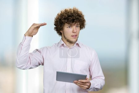 Photo for Portrait of a young man in white shirt holding a tablet pc showing height of something with hand. Male entrepreneur with curly hair giving a speech indoors. - Royalty Free Image